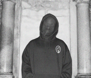 Omni Auspice in a black hoodie with his face covered in black material, framed by two marble pillars. 