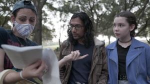 Koura Linda wears a cloth mask and a backwards baseball cap holding a script and talking to an actor with long dark hair in a brown jacket and a young actress with light brown hair in a blue jacket.