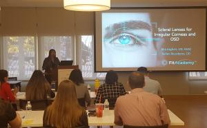Residents sitting at tables in conference room, during a didactic session of FitAcademy.  Screen shows the title "Scleral Lenses for Irregular Corneas and OSD." Dr. Karen Carrasquillo is at podium presenting.
