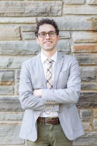 Image of Dr. Nicholas Reed standing in front of a grey stone wall from the waist up wearing glasses and a light grey sports jacket with a plaid tie.