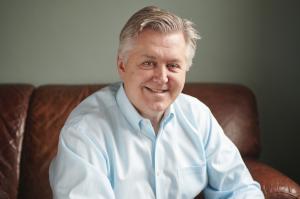 Filmmaker, founder of video production agency Tribe Pictures, and industry thought leader Vern Oakley looks focused facing the camera in a professional headshot. He wears a light blue dress shirt and sits on a brown leather couch.