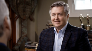 Vern Oakley is smiling, facing NPR journalist David Brancaccio in an interview. Behind Vern, out of focus, are several international film awards.