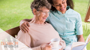 Elderly women sitting with a caregiver aide reading a book on a garden bench.