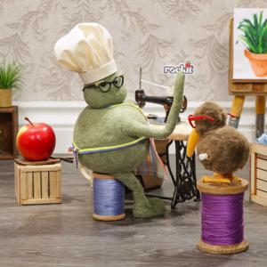The Tiny Chef sits at his sewing machine with a tiny Rockit™ Apple behind him.