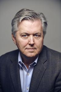 Director Vern Oakley wears a grey suit jacket with a blue shirt and looks sternly at the camera.