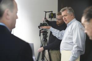 Director Vern Oakley stands between a camera and a white male CEO, offering instructions using hand motions and speaking.