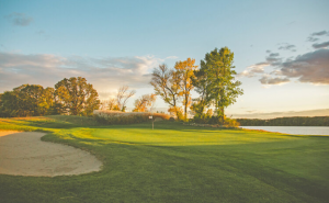 A golf course in the evening, the green is in the center with the white flag standing up, a sand trap is on the left, a lake is on the right, trees in the background, blue sky with two large clouds