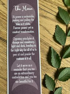 dark purple bookmark with a moon phase design inscribed with a moon inspired poem reading, "It's power is undeniable, pushing and pulling the tides as it orbits. Forever present yet in constant transformation. Opposing principles of change and consistency