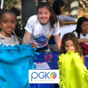 Two children hold up hand-made blankets, next to a volunteer in a Project Giving Kids t-shirt, at a Create the Change volunteer event.