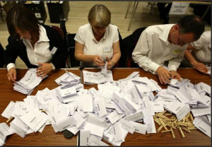 Three workers sit at a table covered with ballots
