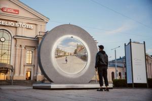 A man looking into a huge outdoor video installation, whose round screen is displaying a different city with a lady walking on the street looking back
