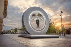 Huge round outdoor art installation in a city with a beautiful skyline providing a portal into another city view with a man looking into the round video screen