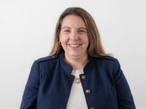 A smiling headshot of Jessica Johnson, chief program and development officer of education northwest. She has on a blue jacket blazer with gold buttons, white crewneck sweater, gold necklace with circular pendant. She has on gold hoop earrings. White backg