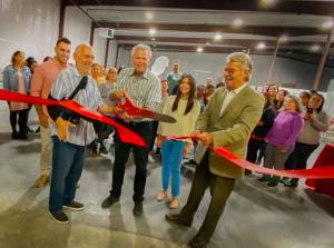 Photograph of the ribbon being cut to commemorate the soft opening it its new building.  The five captioned people cut the giant red ribbon in the foreground, while Coast to Coast employees look on in the background.