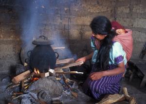 A woman in Guatemala cooks over a traditional open fire, as a baby rests on her back. She is adding wood to the fire as smoke pours into her home.