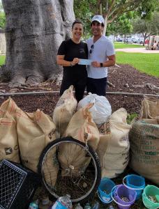 ALT="Dr. Amir Mohit-Kermani, a South Florida chiropractor, presents a donation to Sophie Ringel, executive director of Clean Miami Beach; both stand under a tree with lots of trash from a neighborhood cleanup"