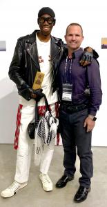 Miss J in all white with a stylish leather jacket poses with the "Gold Bar" Award standing next to Collaboration Artist and Celebrity Photographer James Moritz wearing a black Gucci shirt with bold leather detailed harness