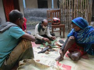 Group of people watching shoemaker construct special shoes. In the foreground, a woman named Sumi watches. Her feet are damaged from leprosy.