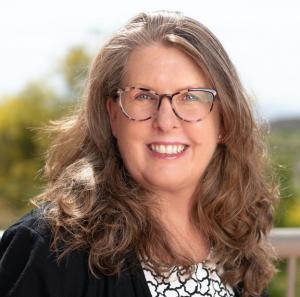 Eileen Bagnall, a White woman with tortoiseshell glasses and shoulder length hair, wearing a black and white blouse under a black blazer.  She outdoors, and smiling.