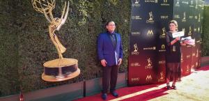 A young man with brown skin and black hair stops for a photograph on a red carpet. He is wearing a tuxedo with a dark blue jacket and dark blue shoes, black formal slacks and a white shirt.  A large statue of an Emmy sits in the background and a young wom