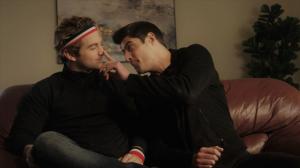 Two young men sit on a couch indoors. One is playfully pressing on the other's nose, while the other looks annoyed.