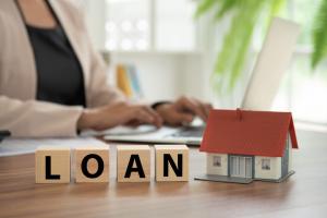 Model home on desk next to wooden blocks spelling the word 'loan' in front of person working on a laptop