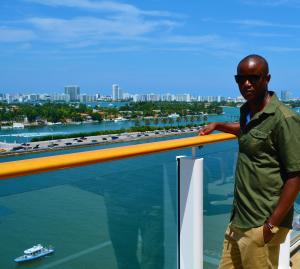 Autrige Dennis wearing a forest green shirt and khaki pants and standing against the background of Miami, Florida.