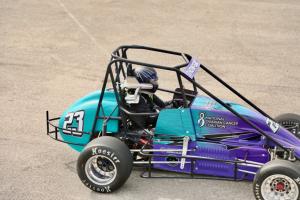 Female driver Ashton Thompson tests out her new race car, driving on the track. The car has a white NOCC logo on the side and is wrapped in teal and purple.