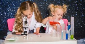 two girls at a table using science equipment. Girl one is using a microscope and girl two is using pouring coloured liquid from beakers and testutbes. This picture promotes girls in stem, stem education, women in stem,