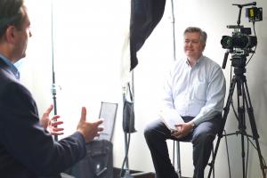 Vern Oakley sits next to a standing camera operator, both looking at a professionally dressed CEO, whose face is not visible in the shot.