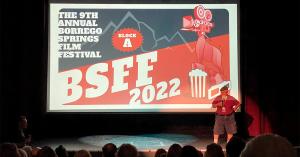 Fred Jee stands on theater stage in front of film screen speaking to festival audience.