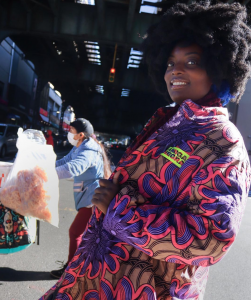 A brown skinned beautiful woman with a large Afro is standing in the middle of the NYC street with a colorfully printed jacket on.