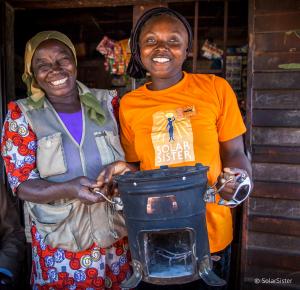 Solar Sister Nanbet Magdalene is standing with one of her customers holding a clean cookstove. They are both smiling broadly.