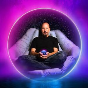Man in black with goatee sits in lounge chair holding blue orb, surrounded in dark pink light.