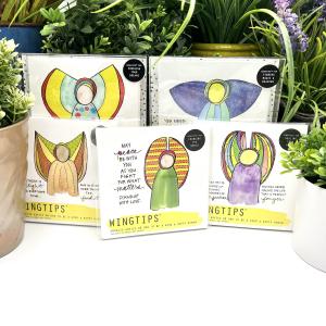 A full collection of 5 WingTips gift sets by Liv Lane are displayed among houseplants on a white shelf. Visible through clear, plastic packaging is top-facing art in each set, with a bright, whimsical angel image and black, handwritten message central to each piece.