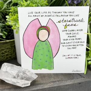 A WingTips print is displayed on a on a white shelf, surrounded by green houseplants and a large clear crystal. The whimsical angel image at the center of the print appears to be wearing a bright green dress with circular blue dots. Pink wings are spread