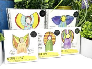 Five WingTips packages are displayed on a white shelf among green houseplants. Top-facing art within each set is visible through clear, plastic packaging, with a bright and colorful angel image central to each piece, surrounded by black, handwritten angel