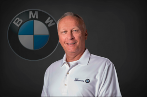 Pictures is Mike Udell Smiling In Front of a BMW logo.