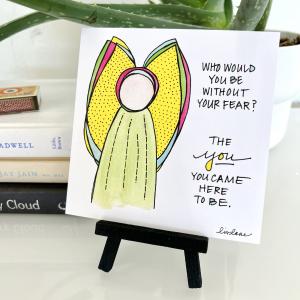 A WingTips print is displayed on a black wooden easel in an office or study. A bright, whimsical angel watercolor sketch is at the left of the print. Handwriting to the right reads, "Who would you be without your fear? The you you came here to be. Signed, Liv Lane