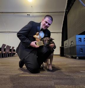 a man in a tuxedo kneels next to a German shepherd, holding a gold trophy