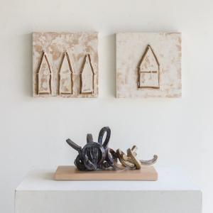 Two clay, plaster,  tan paints that have beam pegs arranged to depict a simplistic houses hung side by side on the wall. Below them sitting on a pedestal is a blue ceramic twisted hoop sitting on a light brown wooden block.