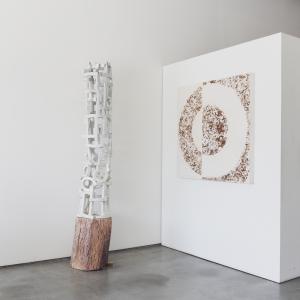 78 inch tall white ceramic tower set on a wooden stand. This tower is standing next to a plaster, paint, clay artwork with a large circle made of wooden chips plastered into the art hung on the wall.
