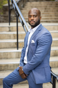 A photo of author James Pierre in a suit on stairs looking at the camera.