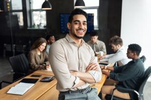 Portrait of confident smiling bearded businessman sitting leaning on desk in office, posing with folded arms and looking at camera, colleagues working in blurred background