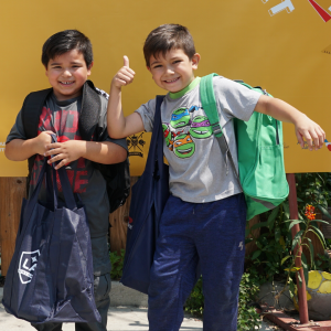 Two APCH members stand in front of a yellow back to school banner with their backpacks. One member gives a thumbs up.