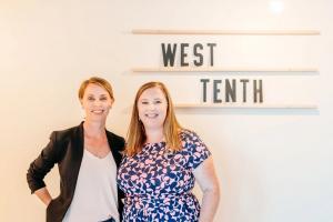 CEO Lyn Johnson and COO Sara Sparhawk stand smiling side-by-side in front of a hand-made sign that reads "WEST TENTH"