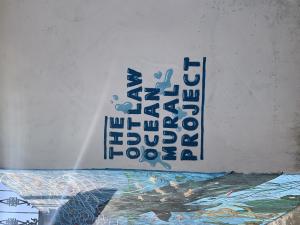 White wall with the words The Outlaw Ocean Mural Project painted in black and clear blue water drops painted around it