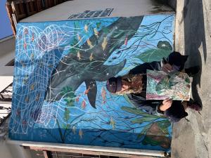Artist Claudia Tostes, wearing black, is in crouching position. She holds a drawing with a draft of the mural. She is in front of a mural of the sea, showcasing plants and corals on the bottom in different shades of green and red, floating over it is a la