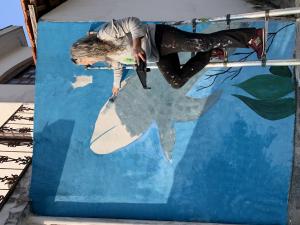 Artist Claudia Tostes on a stepladder in the middle of painting a whale on a wall painted blue.