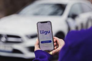 Woman is holding a phone with Liigu app's front page open as if about to book a rental. In the background there is a white car with purple Liigu letters spelling the company's name.